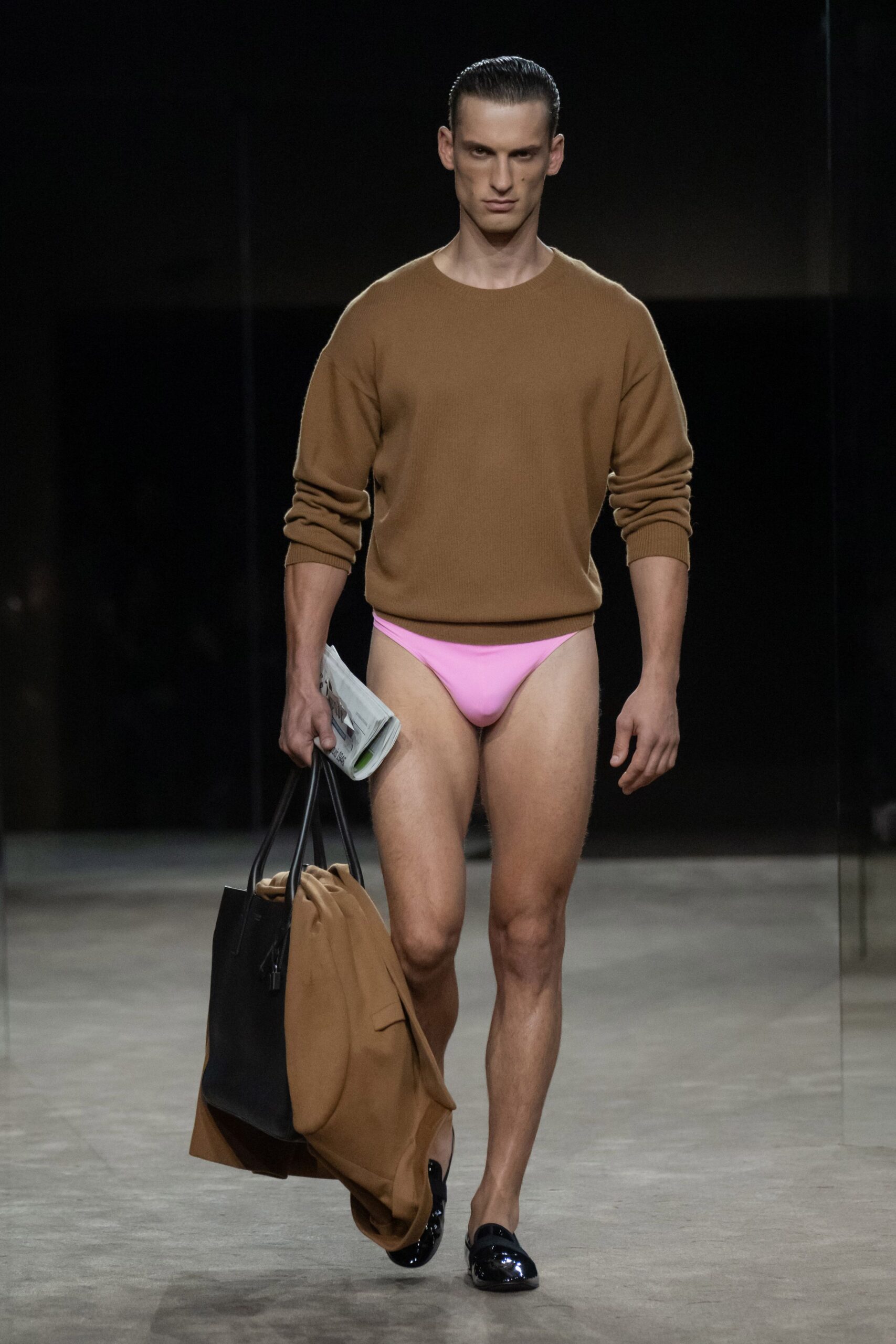 Camel fine-gauge crewneck sweater with a close ribbed hem. Bubblegum-pink briefs cut high on the hip bone. Folded newspaper tucked under one arm. Black leather tote carrying a camel coat slung through it. Black patent loafers catching white glare at the toe. Early paper run, still half dressed, coffee not yet in hand.