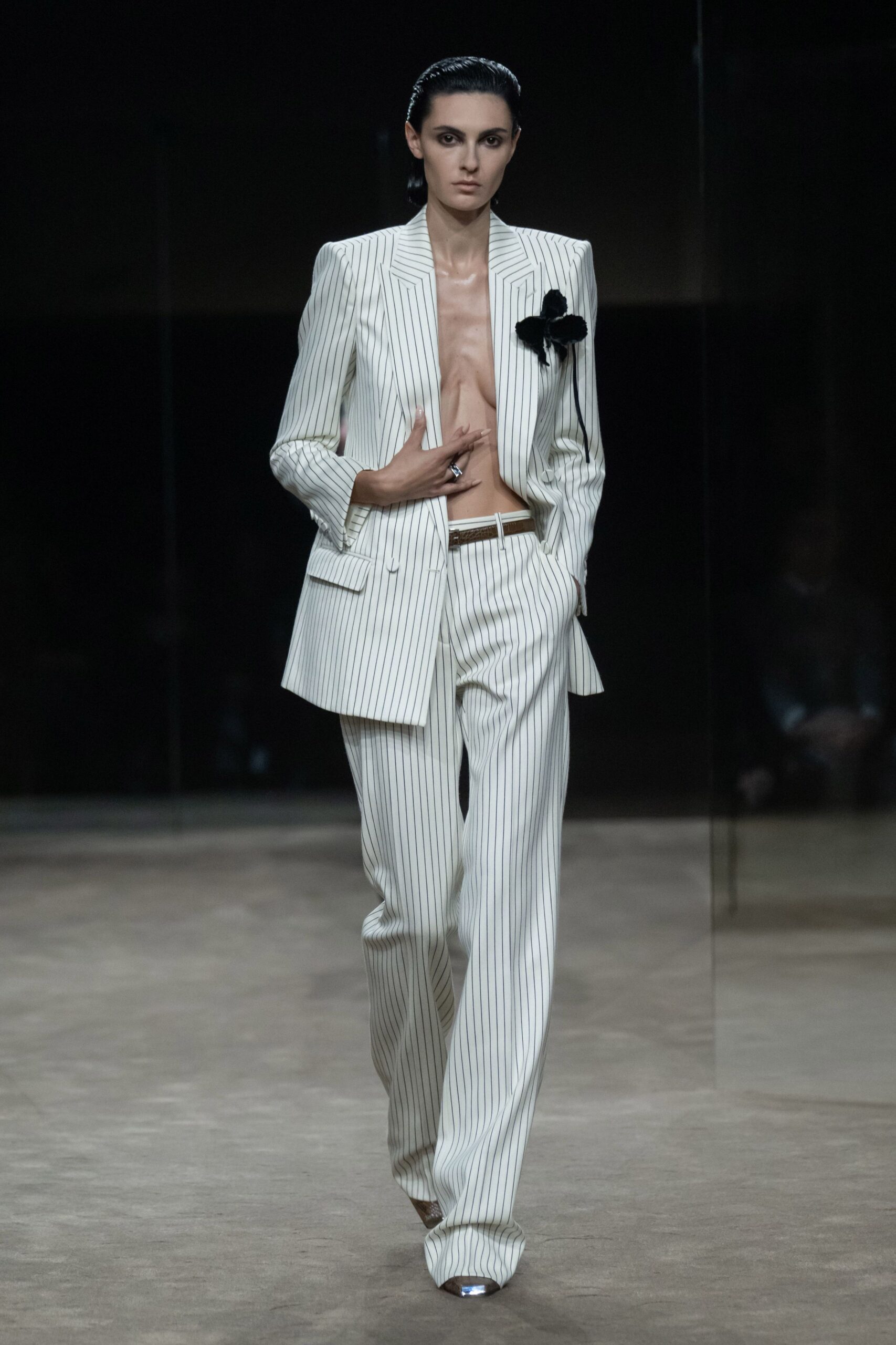 Ivory pinstriped blazer cut with sharp shoulders and a black pompom corsage. Matching pinstriped trousers pooling over the shoe, brown belt just showing. Metal-capped pointed pumps. For crossing a hotel lobby before anyone has ordered breakfast.