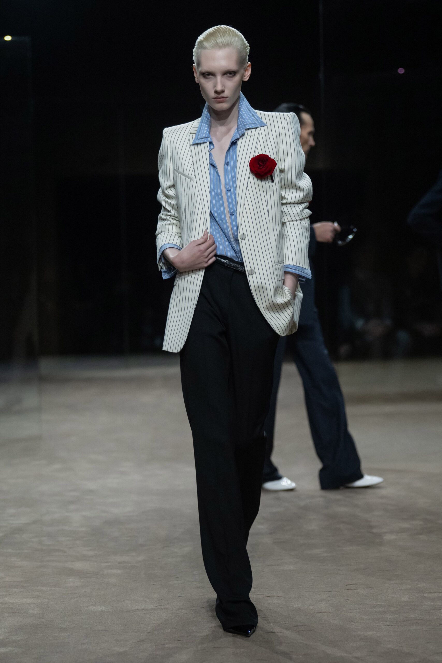 Cream pinstriped blazer with pagoda shoulders and a red rose pinned high. Powder-blue banker stripe shirt, collar spread wide and cuffs turned back. Black tailored trousers breaking softly over a pointed shoe. Glossy black pointed toes. Crossing a hotel lobby before breakfast, still carrying last night's argument.