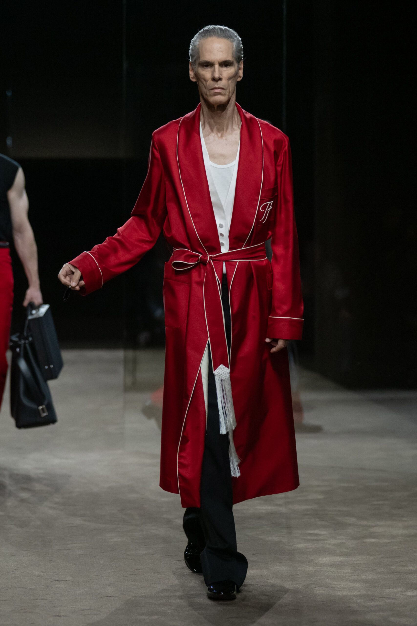 Crimson satin robe coat with white piping and a long tasselled tie. White ribbed tank underlayer, scooped low at the chest. Black trousers falling straight from beneath the robe. Black patent shoes with a blunt square toe. Hotel corridor at dawn, carrying the room key after no sleep.