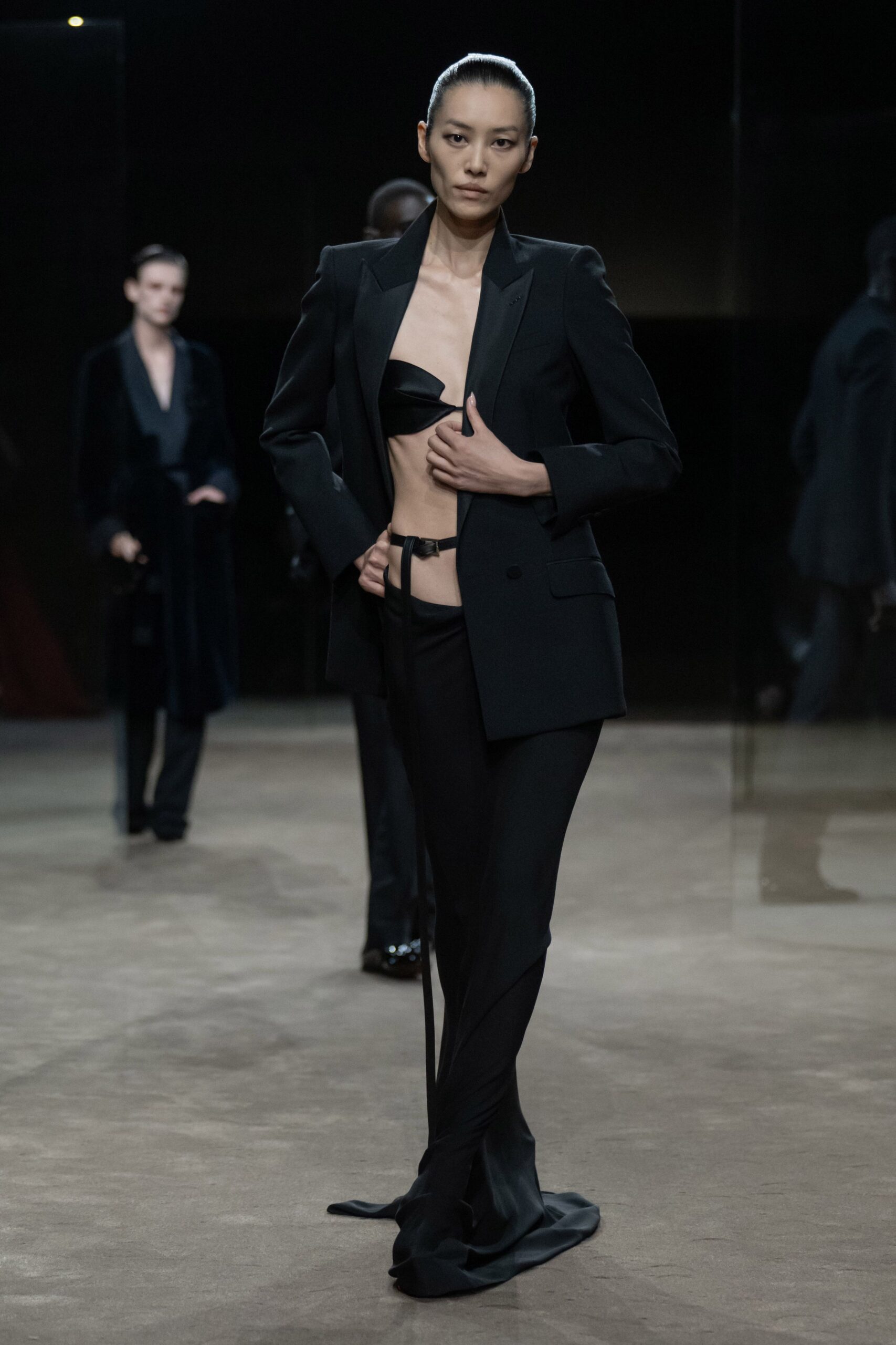 Black tailored blazer with peaked satin lapels and a low double button. Tiny black satin bandeau, one side pulled to a narrow point. Black fluid trousers cut low on the hip with a trailing hem. Thin black belt, loose tail hanging past the thigh. Whatever you wear home when the hotel bar piano has stopped.