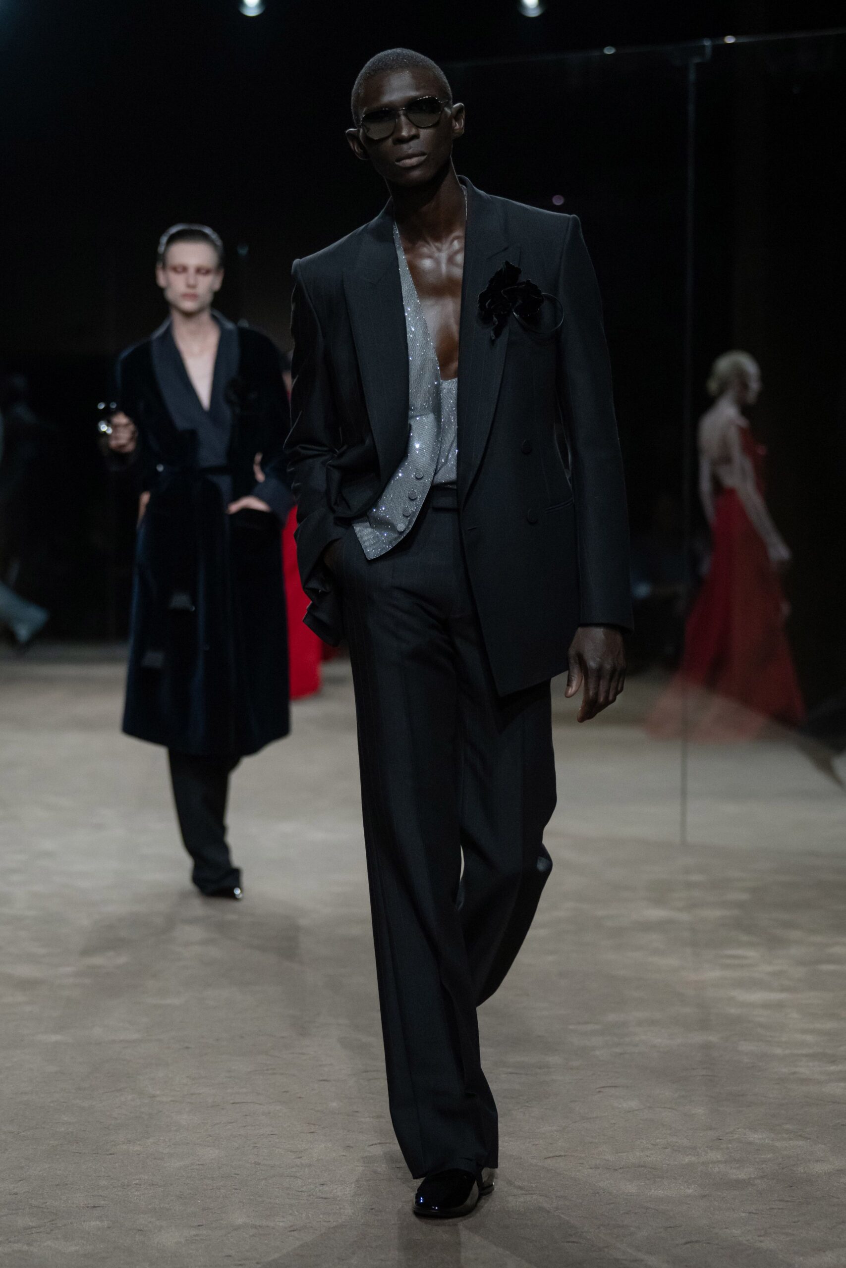 Dark square lenses in a thin metal frame. Black double-breasted blazer with a velvet flower at the chest. Silver glitter waistcoat cut plunging low, four covered buttons. Black tailored trousers falling in a long clean break. Black patent square toes. Walking into a hotel bar just after the wedding has ended.