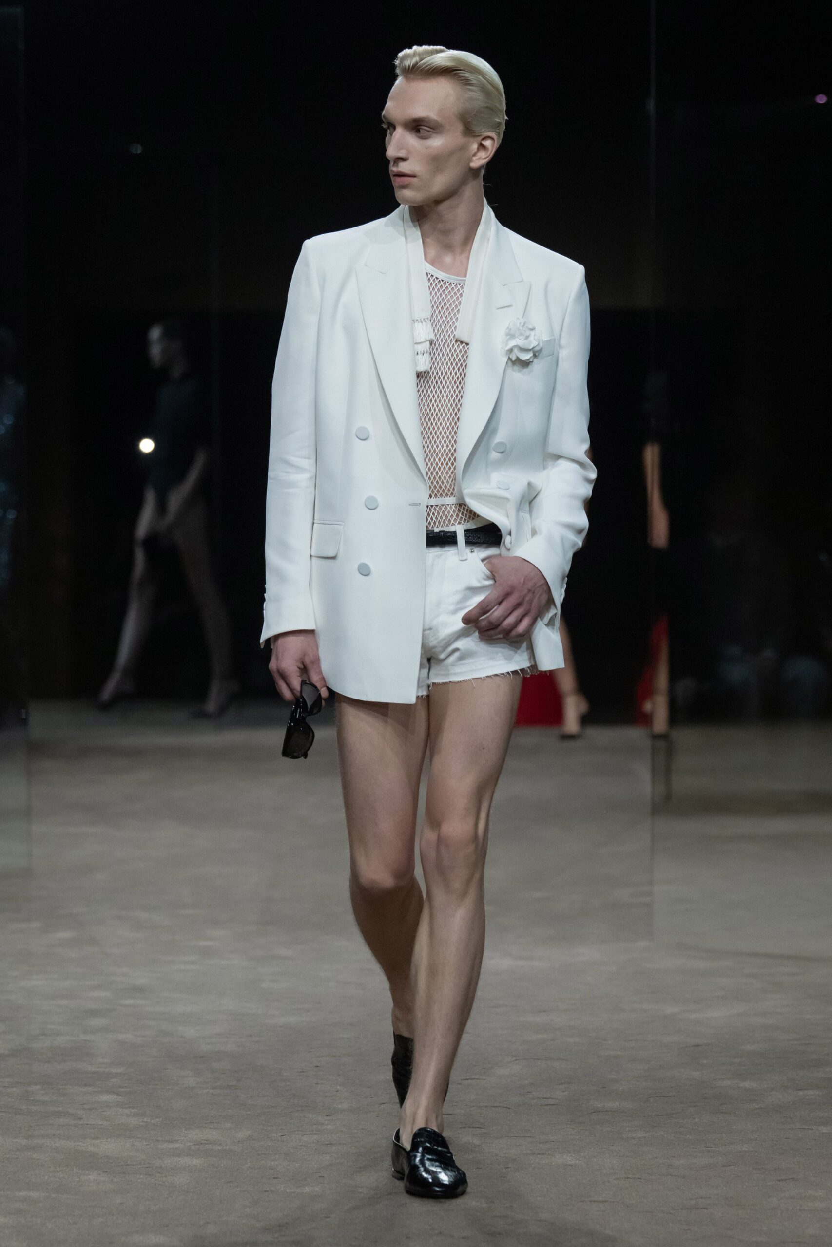 White double-breasted blazer with peaked lapels and a pinned fabric flower. White open-net tank layered over ribbed bands at the chest. White cutoff shorts with a frayed hem and high black belt. Black patent loafers. Black rectangular sunglasses in hand. Walking into a hotel lobby still dressed for the pool party upstairs.