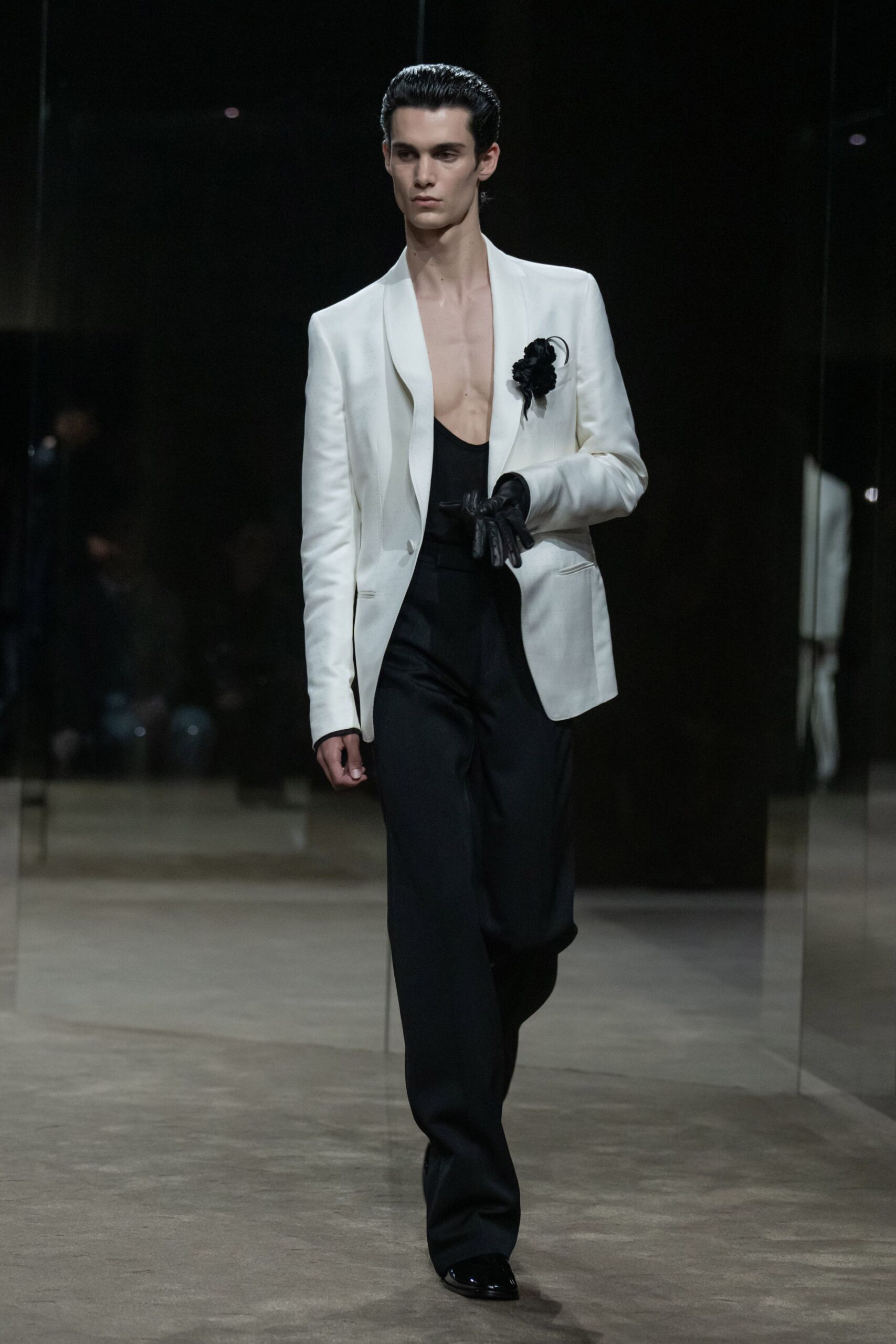 Ivory shawl-collar blazer worn bare, a black flower pinned high. Black scoop tank cut low through the chest. High-waisted black trousers falling in a long, clean drape. Black leather gloves carried in one hand. Patent black square toes. Crossing a hotel lobby after midnight with someone else's room key.