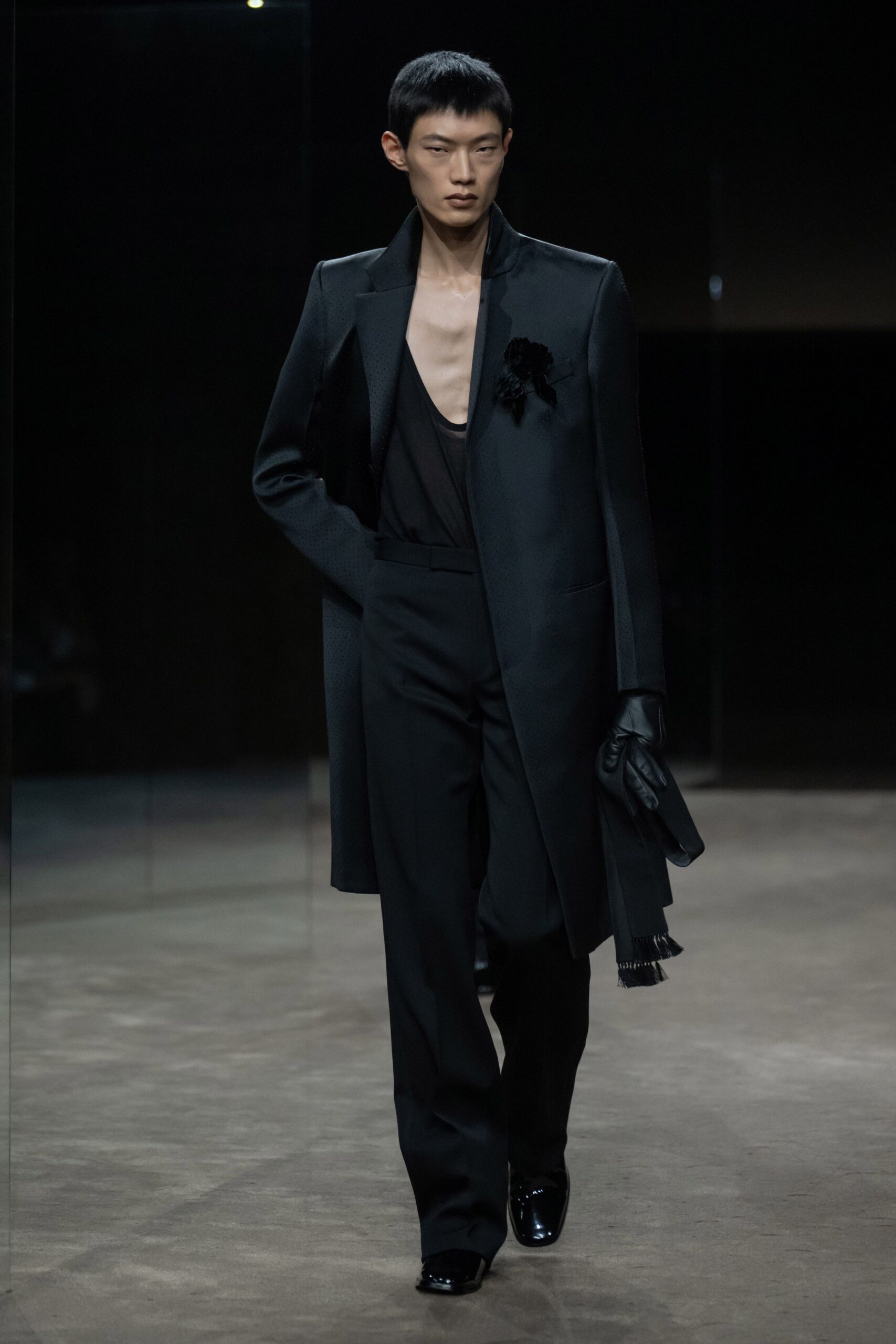 Long black coat with satin-faced lapels and a frayed scarf hem. Deep scoop black jersey tank, cut almost to the sternum. Black tailored trousers with a high rise and soft break. Black leather gloves. Square-toe patent shoes. Black flower pinned at the chest. For arriving at a memorial after everyone else has already gone quiet.