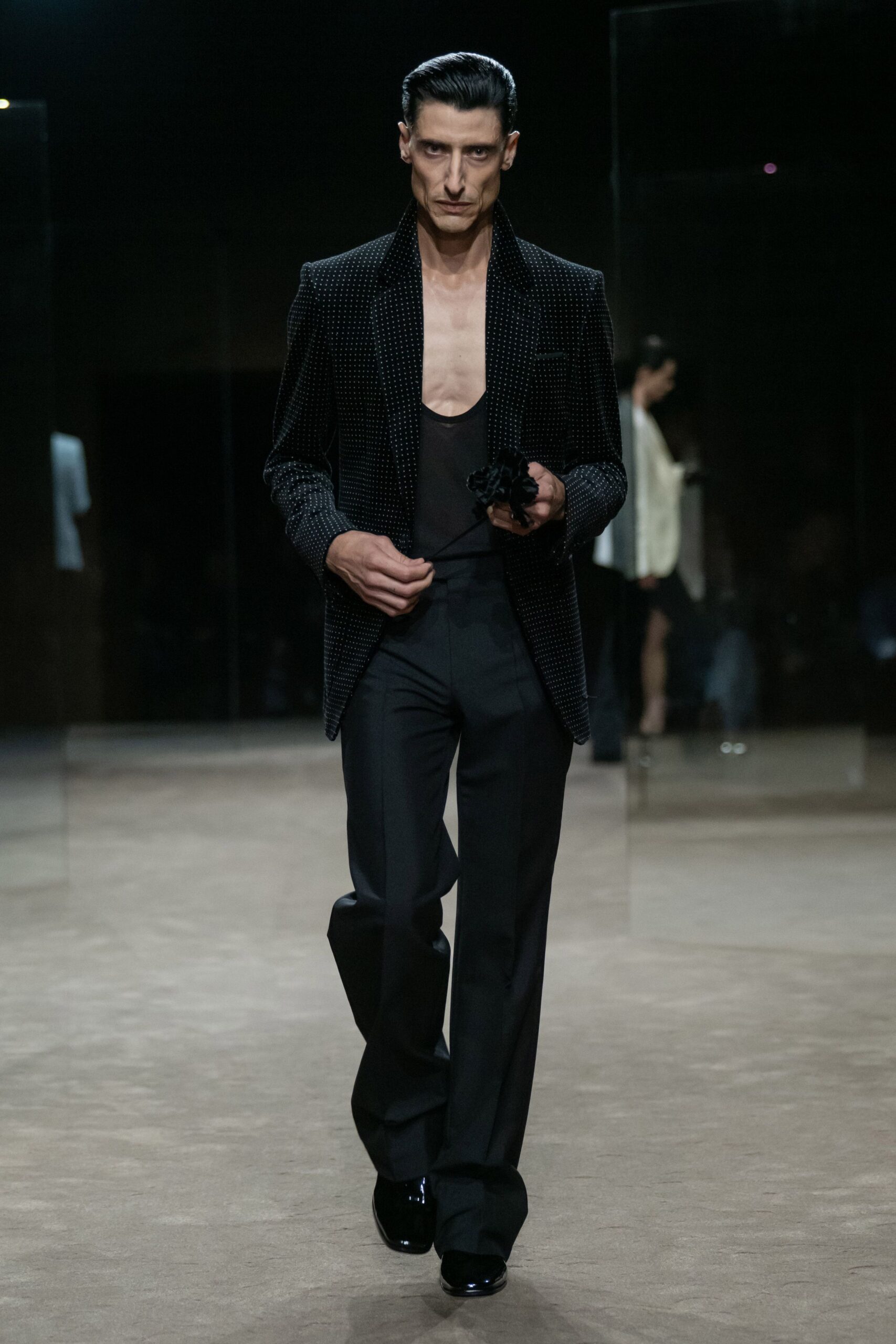 Black blazer dusted with tiny silver studs, collar spread wide. Low black tank scooped almost to the sternum. Black tailored trousers with a long break over the shoe. Crushed black rosette gripped in one hand by a thin stem. Glossy black square-toe shoes. Crossing a hotel atrium after midnight, boutonniere already pulled off.
