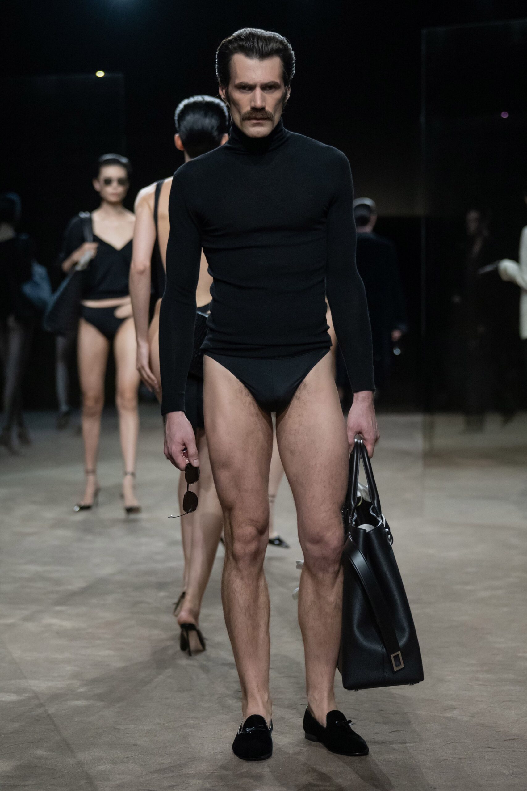 Black fine-gauge knit turtleneck stretched close through the torso. High-cut black briefs with a spare swimwear finish. Small dark aviators dangling from one hand. Oversized black leather tote with a square metal tab. Black velvet slippers with a tiny bow at the vamp. For answering the villa door before anyone else is dressed.