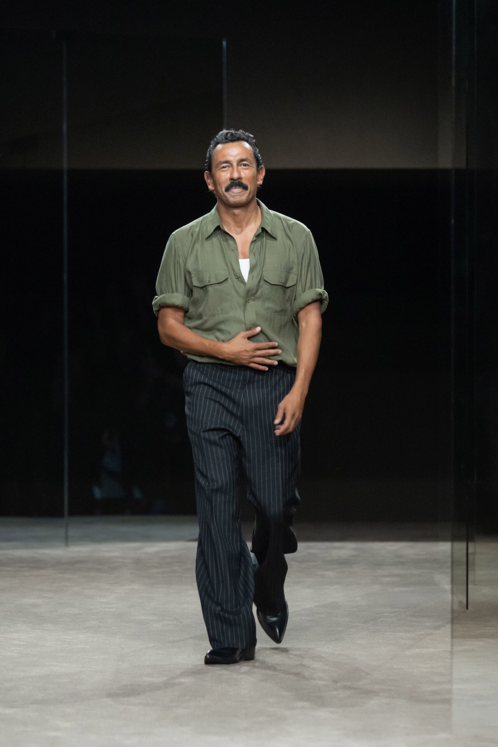 Olive camp-collar shirt in fluid twill, sleeves rolled thick at the bicep. White ribbed tank peeking through the open placket. Charcoal pinstripe trousers with a long, easy break over the shoe. Black leather pointed boots. For the walk home after dinner, still warm and carrying cigarettes.