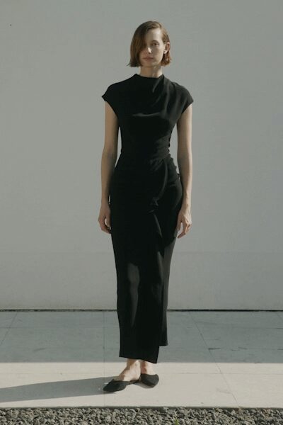 Black jersey column dress with cap sleeves and a soft draped neck. Black pointed flats cut low across the foot. Midmorning in a bare apartment, the curtains still not hung.