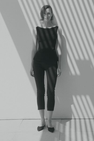 Black sleeveless top with a wide bateau neckline and sharp shoulder points. Black fitted capris cropped above the ankle, smooth and close through the calf. Dark pointed flats with low-cut vamps. Midmorning sun striping the hallway outside a quiet apartment.