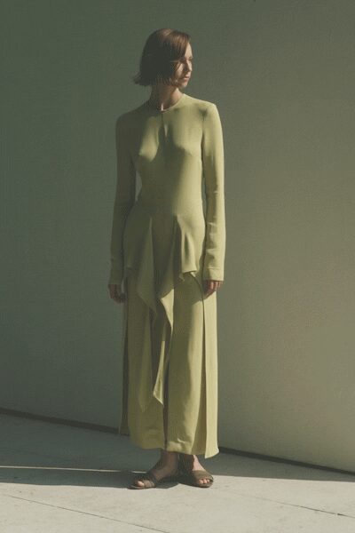 Celery jersey column dress with long sleeves and a twisted drape falling from the hip. Flat brown leather sandals, bare and plain against the pooled hem. Afternoon heat in a rented house, doors open to a chalky terrace.