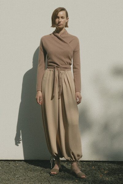 Taupe ribbed knit top with a folded funnel neck crossing off-center. Sand paperbag trousers, softly gathered under a self-tie waist. Nude ankle-strap sandals with slim crossed toe bands. Standing in the courtyard after watering the plants, sleeves still cool from shade.