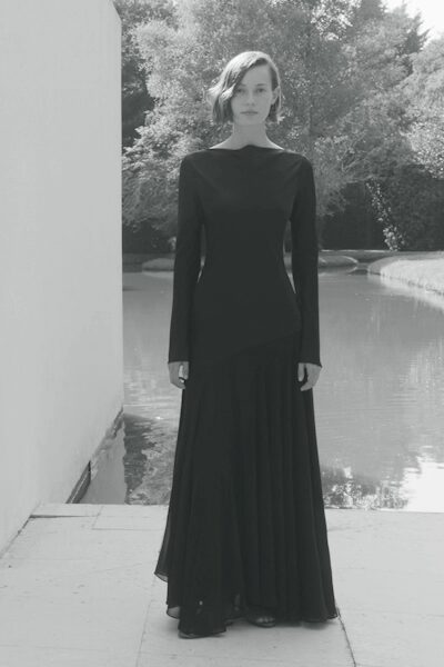 Black column gown with a bateau neck and long sleeves flaring past the wrists. Sheer godets dropped from a slanted hip seam, loosening into the floor. By the reflecting pool after sunset, when the event has thinned out.