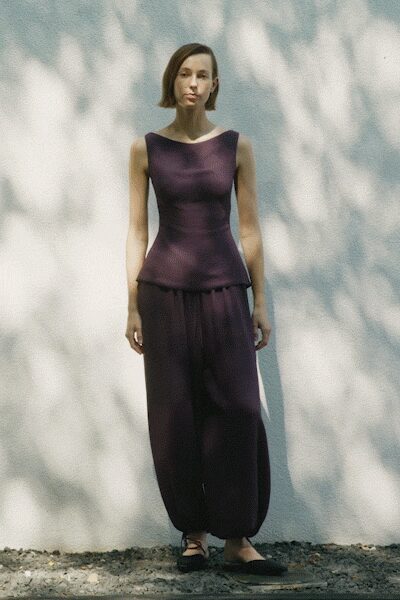Plum stretch-knit top, boat neck and peplum hem skimming over the hips. Matching plum trousers cut full and gathered tight at the ankle. Black wrap-tie flats crossing the instep. Standing in yard shade while the laundry line throws branches on the wall.