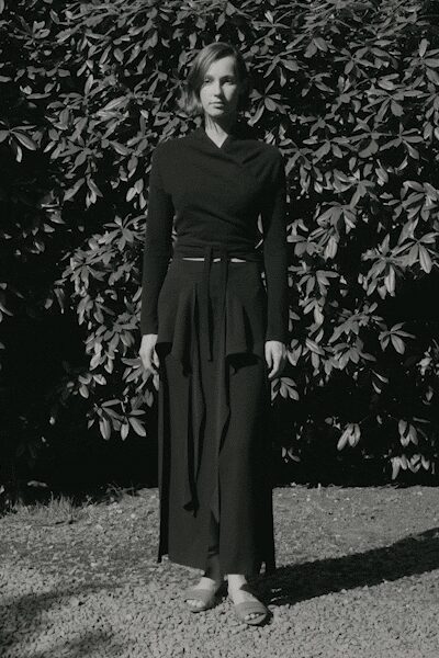 Black wrap top in fine jersey, crossover pulled to a soft shawl collar. Narrow self-tie belt, one long end dropping past the hip. Floor-length black skirt with deep knife pleats opening from the waist. Pale open-toe sandals with a broad front strap. Standing on gravel in hard noon sun, hedge throwing leaf-shadows everywhere.