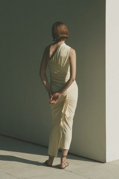 Pale one-shoulder jersey dress, tightly ruched through the back and hip. Thin black lace-up sandals winding around the ankle. Late afternoon by a whitewashed wall, heat still rising off the pavement.