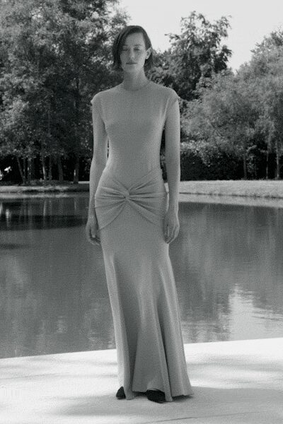 Sleeveless column gown in fine rib knit, twisted into a gathered knot at the hips. Beside a still pond before lunch, when the heat has flattened everything.