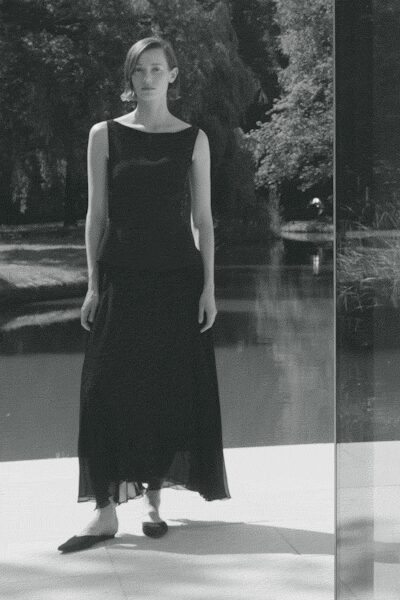 Black sleeveless dress with a wide bateau neck and softly dropped waist. Sheer gauze hem drifting below in an uneven ankle-length tier. Pointed dark flats with low-cut vamps. By the ornamental pond before guests drift out for lunch.