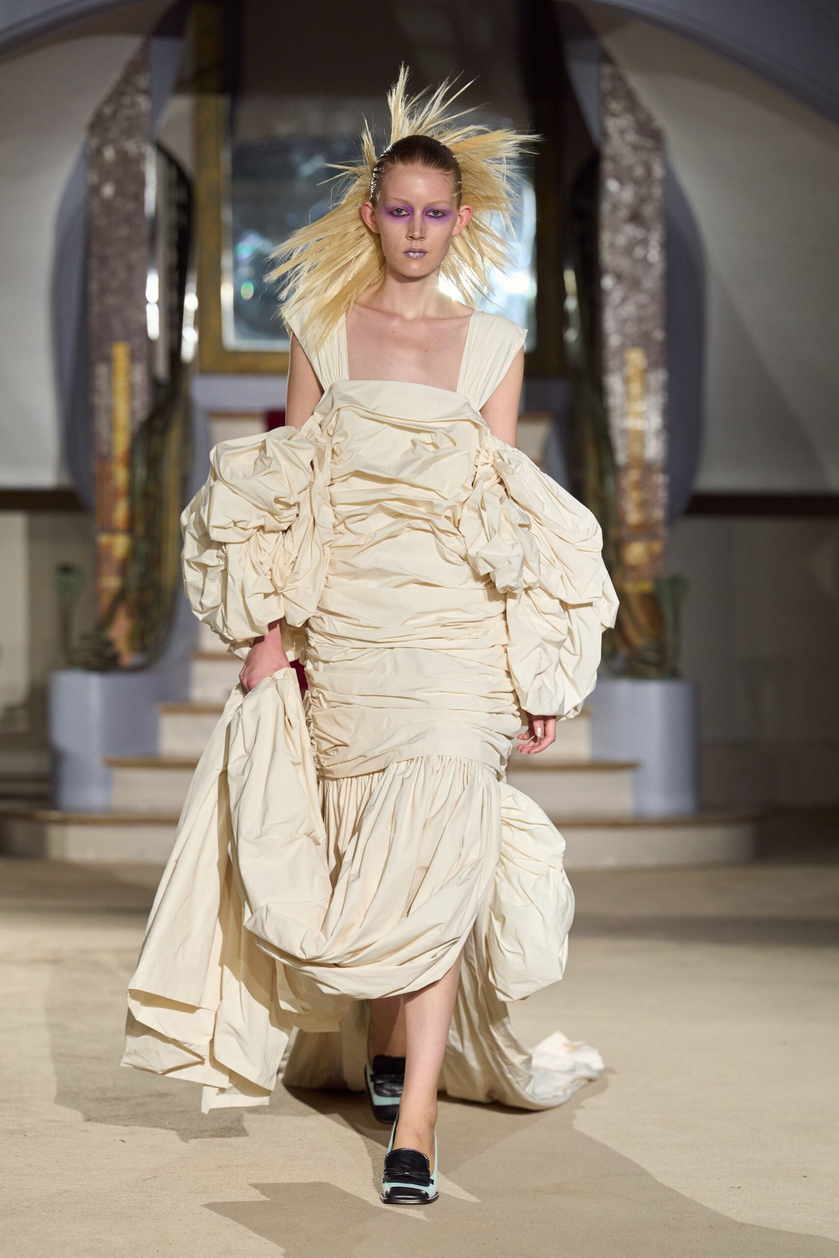 Cream taffeta dress ruched through the torso with ballooned dropped sleeves. Gathered overskirt bunched in one hand, trailing behind in crushed folds. Square-toe pumps with black vamps and pale mint sides. Descending a hotel staircase after sleeping in yesterday's formal clothes.