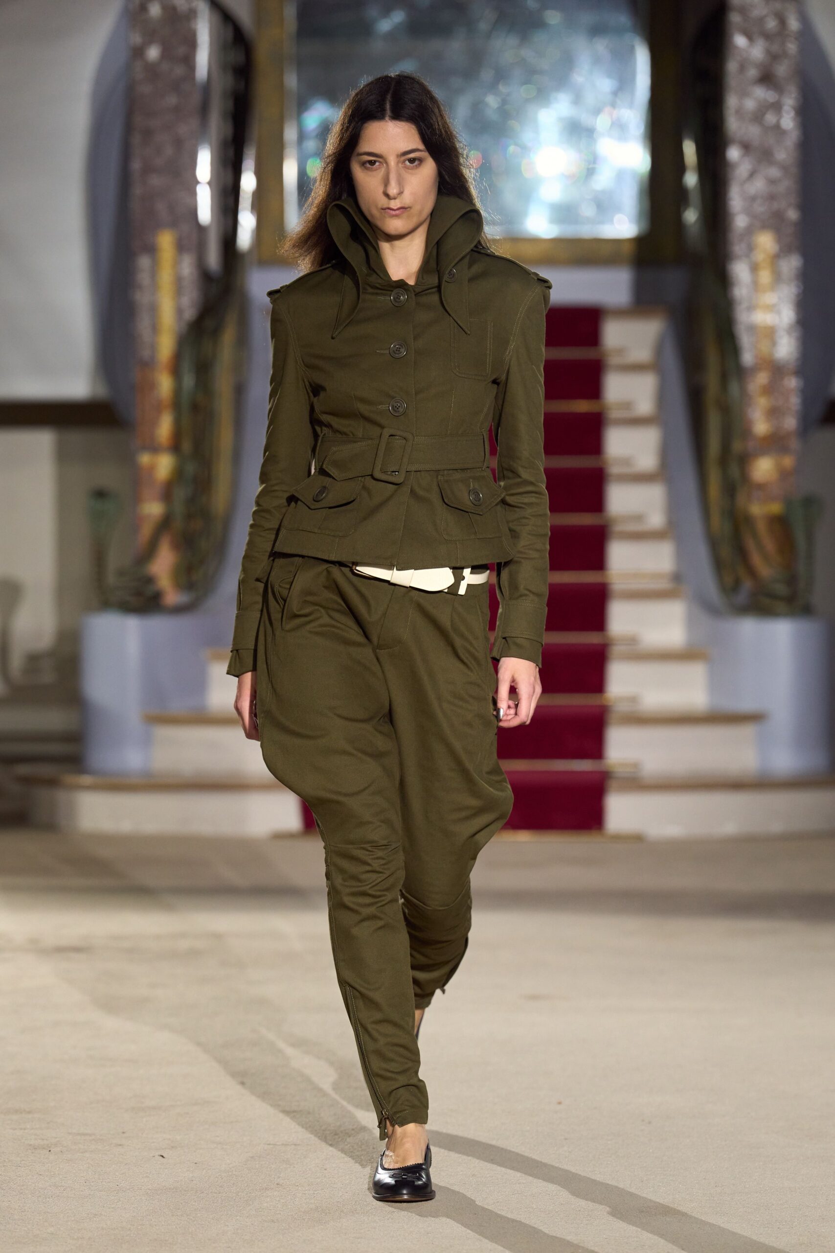 Olive cotton drill jacket with a high sculpted stand collar. Self-belt with a square covered buckle cinching the waist. Matching tapered trousers, zip vents opening at the ankles. White belt peeking crookedly beneath the jacket hem. Black-and-silver cap-toe flats. For checking into a provincial hotel just before the rain starts.