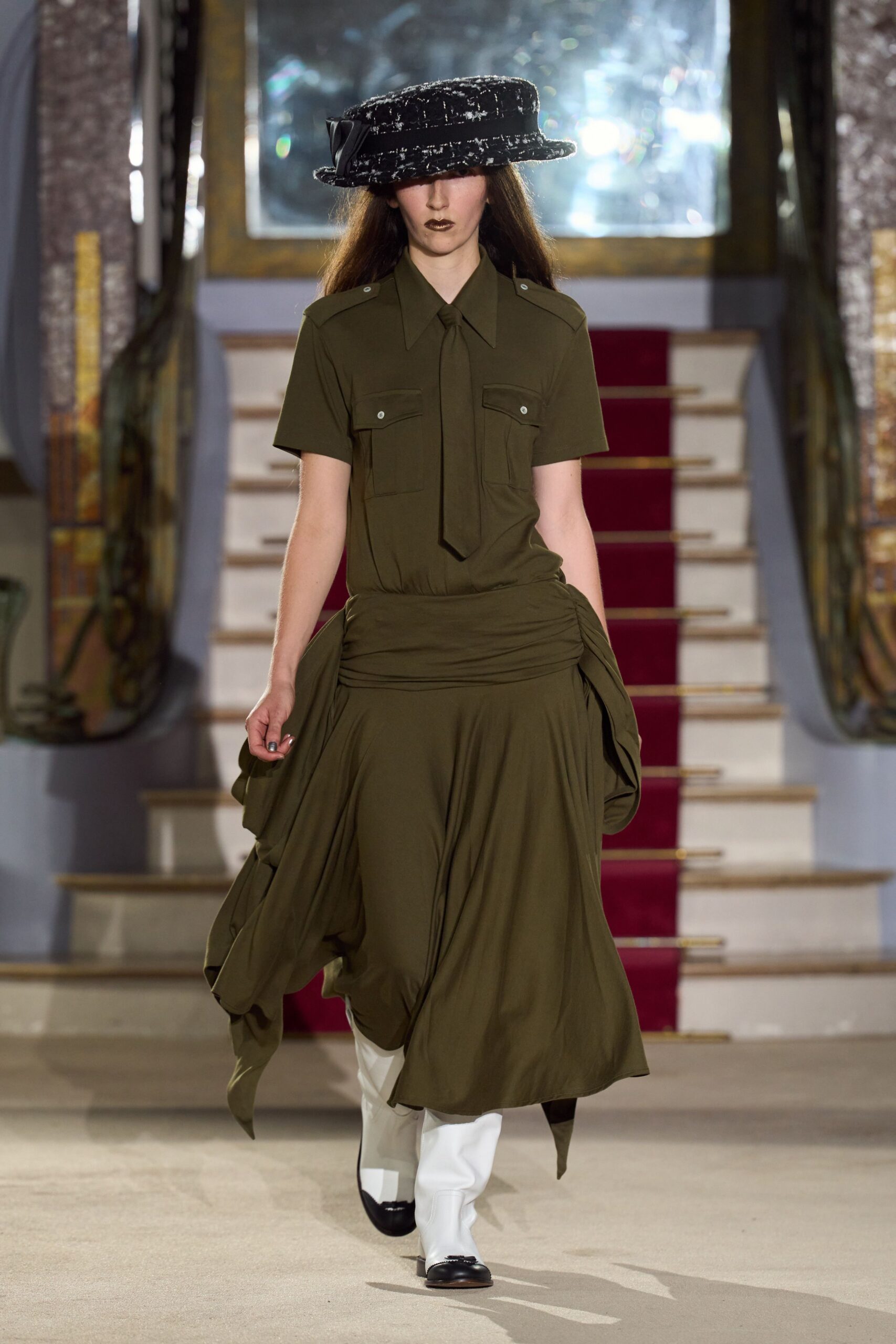 Black boucle boater, brim crusted with white flecks and a side bow. Olive crepe shirt dress with epaulettes, flap pockets and a narrow tie. White knee boots with black dipped toes. Crossing a municipal hall after rain, still carrying the meeting in your shoulders.
