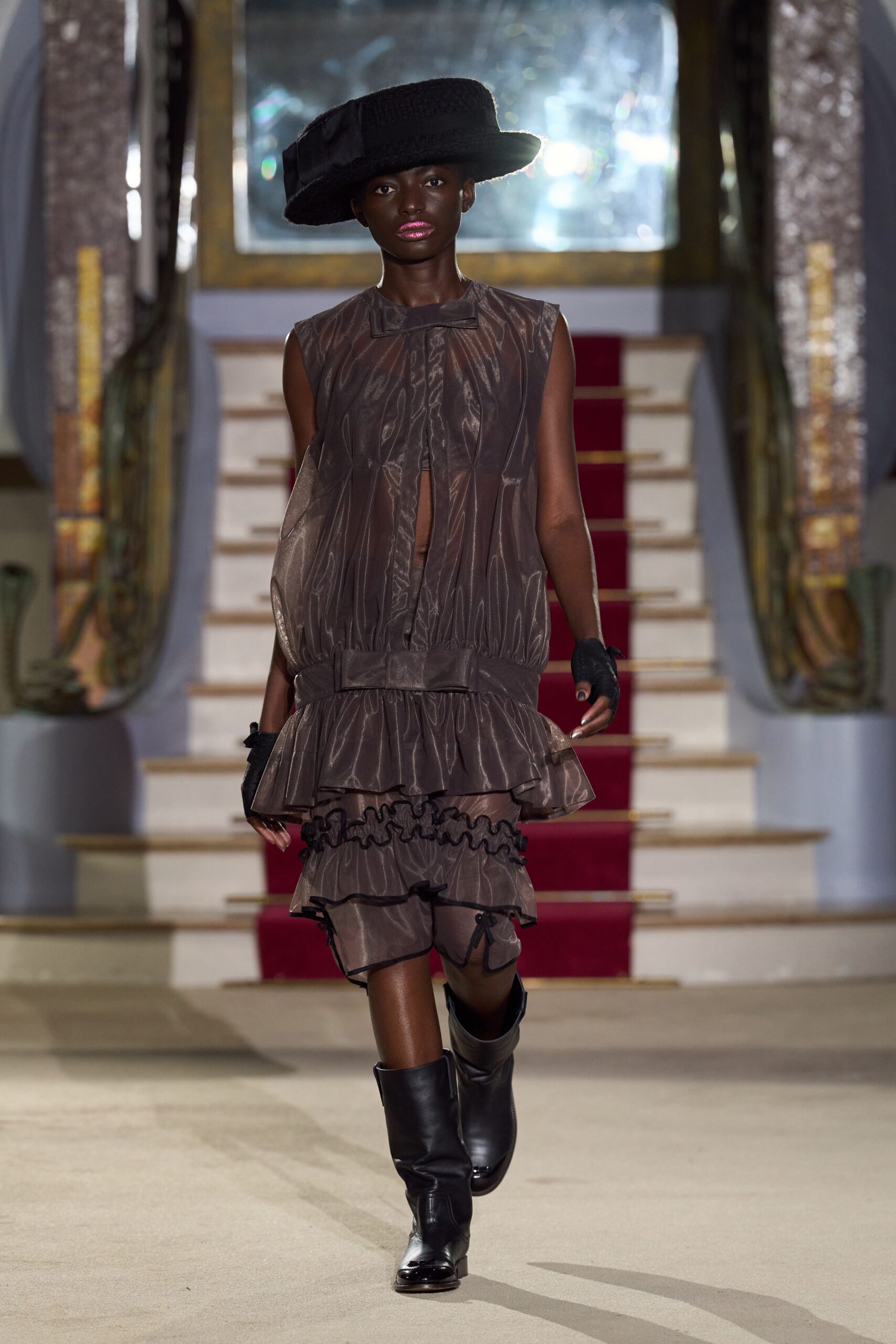 Wide black felt hat tipped low, brim squared off at one side. Smoke-brown sheer dress, gathered into a dropped waistband with ruffled tiers. Black fingerless gloves. Slouchy black boots with glossy square toes. Crossing a hotel lobby before breakfast, lipstick still louder than the room.