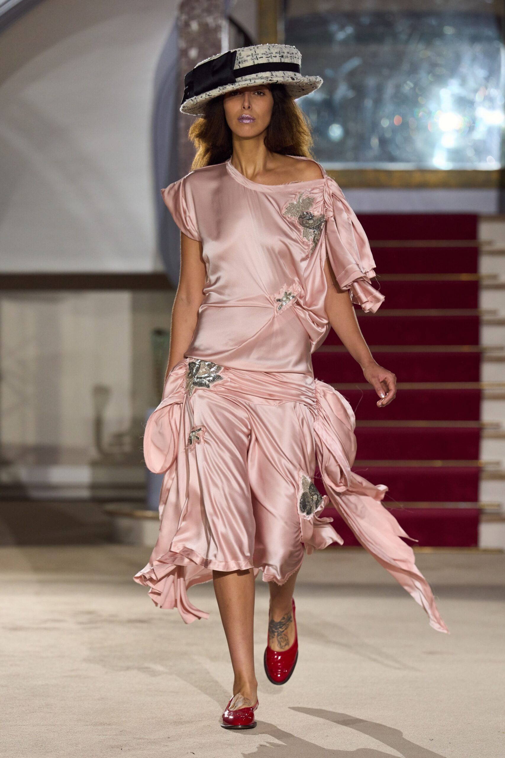 White tweed boater with a wide black bow and script flecks. Blush satin dress, one shoulder slipping low, ruched into silver appliques. Cherry patent mules with pale tufts at the vamp. Descending the hotel staircase after the wedding band has packed up.