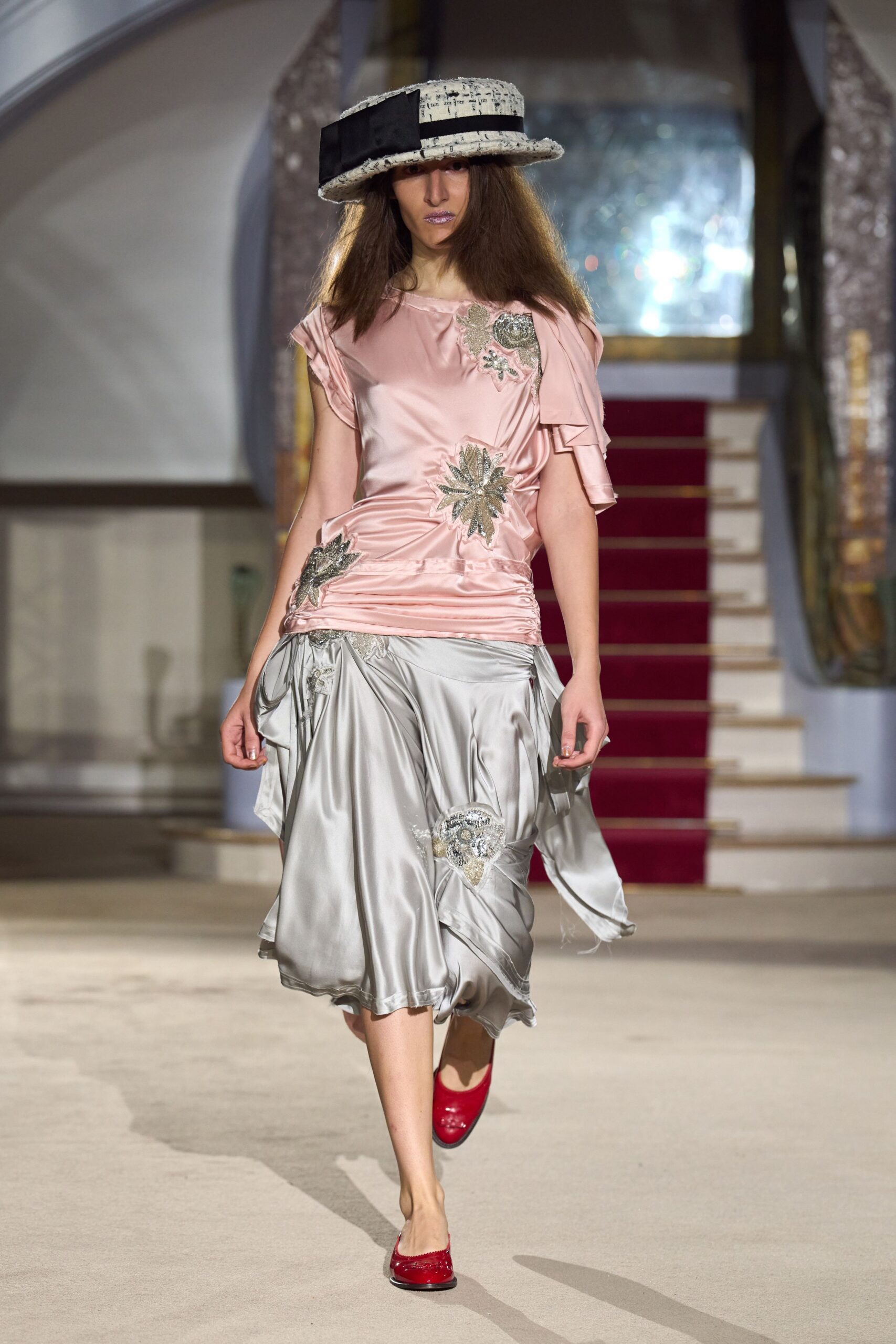 Cream bouclé cloche with a blunt black grosgrain band and square bow. Blush satin top, side-ruched and patched with silver beaded florals. Pearl-grey satin skirt, handkerchief panels swinging off the hip. Cherry patent flats. Crossing a hotel ballroom carpet before the wedding guests are let in.