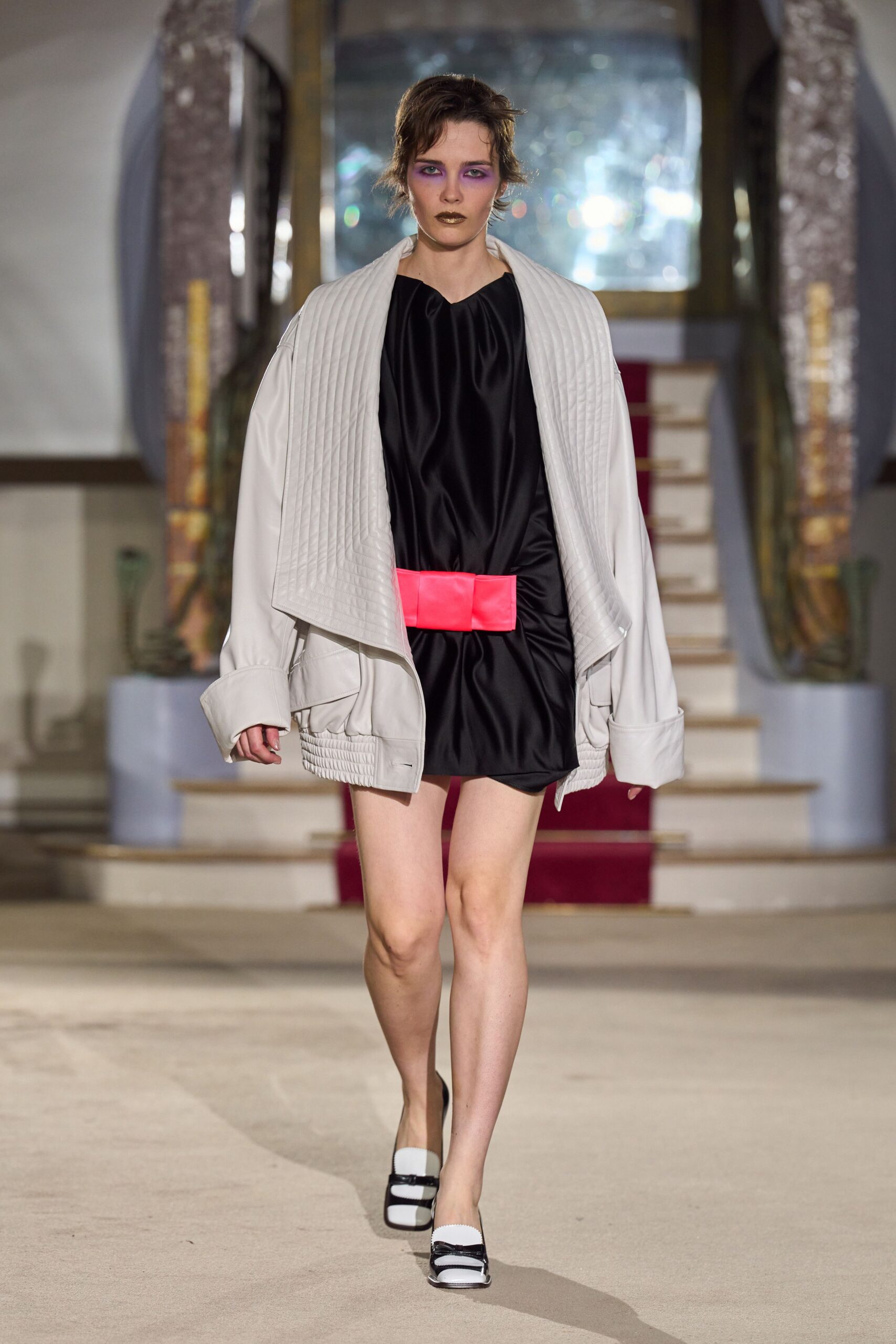 Ivory leather jacket with channel-quilted lapels and turned-back cuffs. Black satin mini dress, bloused soft above a neon coral belt. Wide neon coral belt, folded into blunt padded panels. Black-and-white square-toe flats with glossy strap bars. Coming downstairs in a hotel atrium after missing the wedding breakfast.