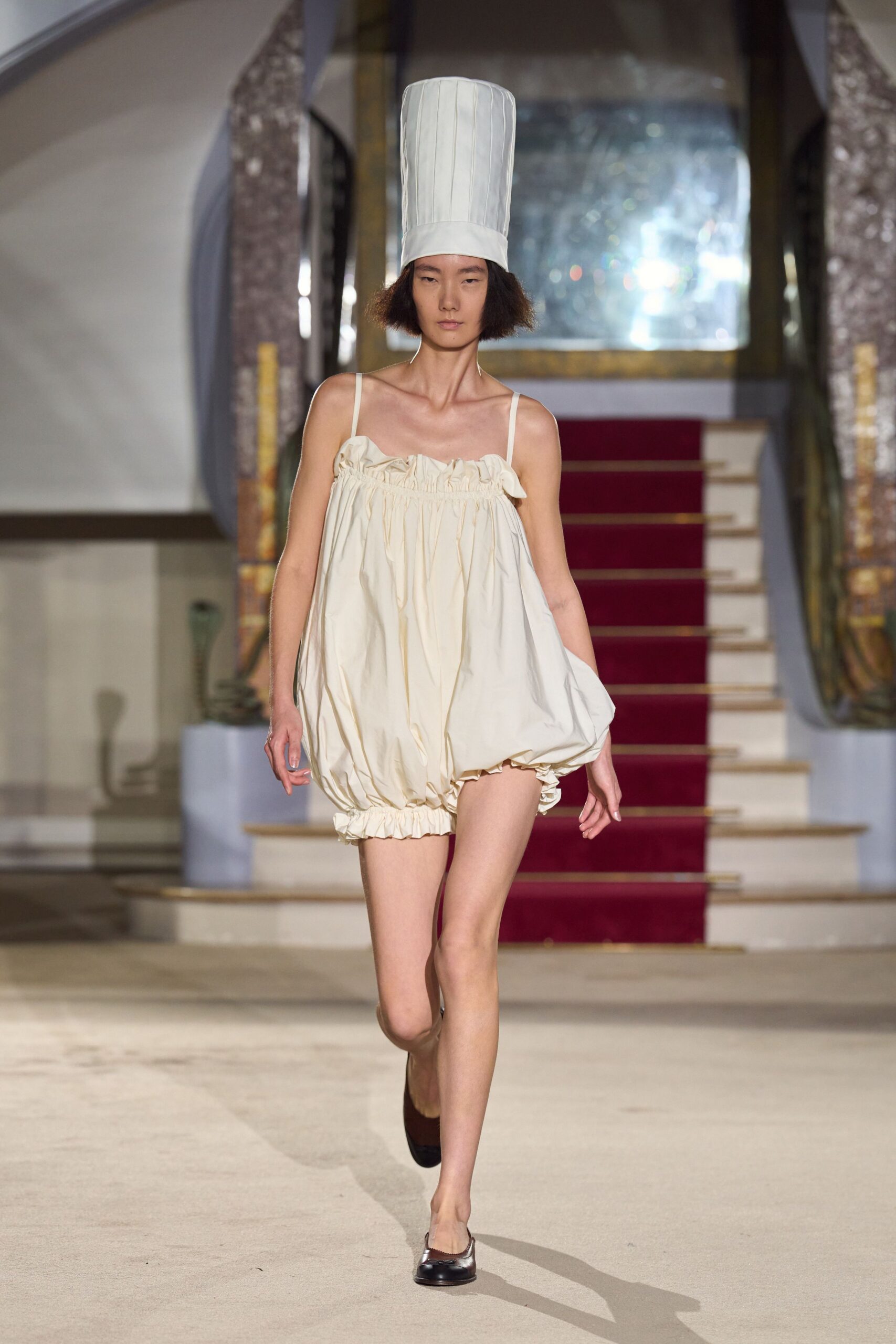 Tall white toque in crisp pleated cotton. Cream poplin bubble dress, ruched neckline and elastic hem puffing outward. Brown square-toe flats with a dark capped front. Crossing a hotel lobby after midnight still dressed from the kitchen prank.