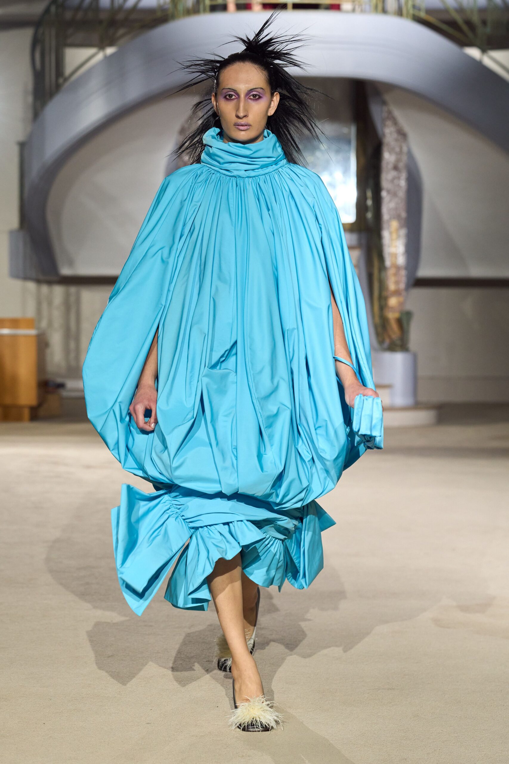 Turquoise taffeta cocoon dress, densely gathered at the neck and ballooned low. Matching folded pouch clutched in one hand. Black satin mules tipped with a pale feather tuft. Crossing a hotel atrium before the fountain switches on for noon.