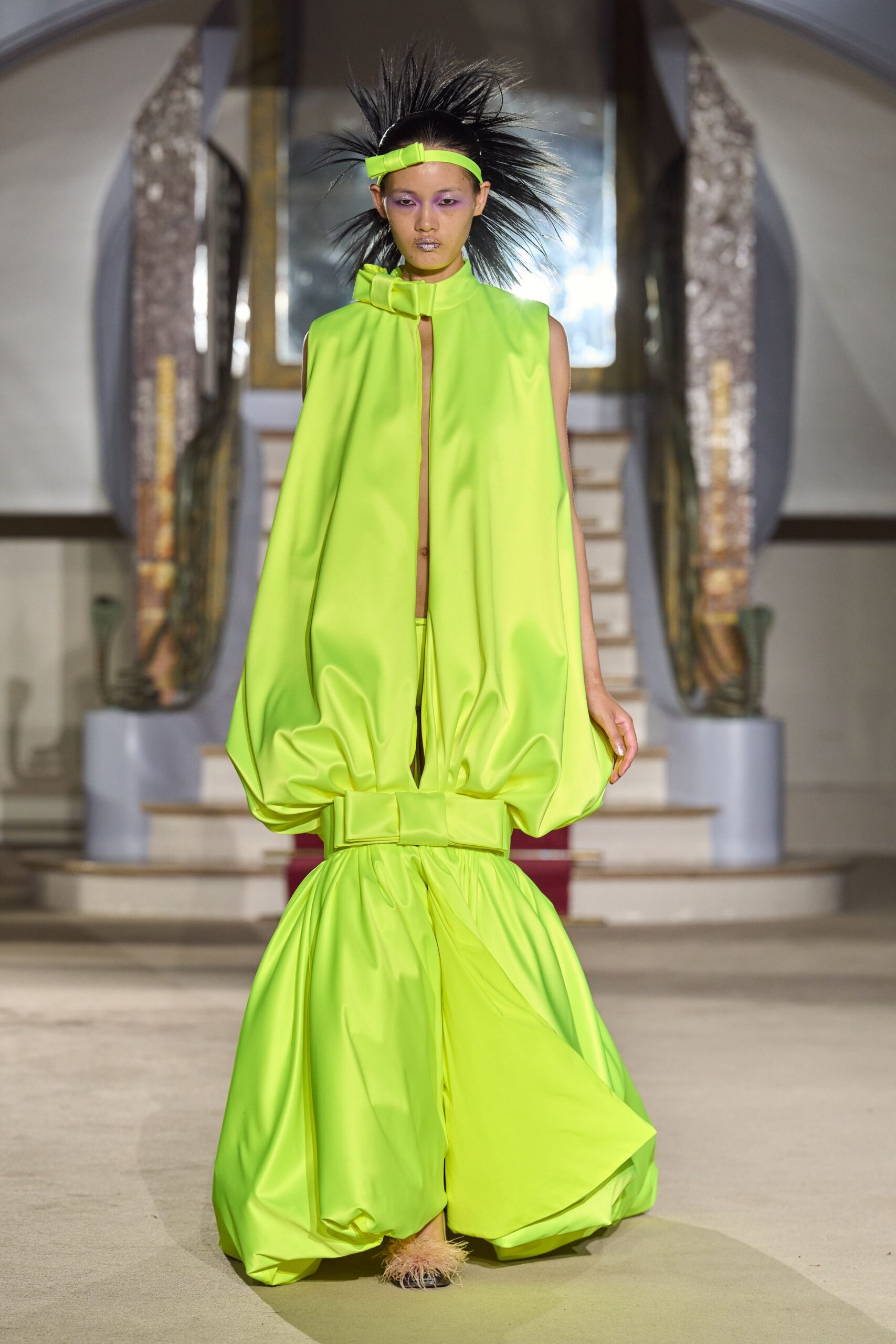 Neon chartreuse band with a squared bow at the temple. High-necked satin cape top, front split open and ballooned at the hem. Matching wide trousers in pooled satin, cinched low with an oversized bow. Barely seen sandals with peach marabou at the toe. Descending a hotel staircase as if the lobby were lit for a game show.