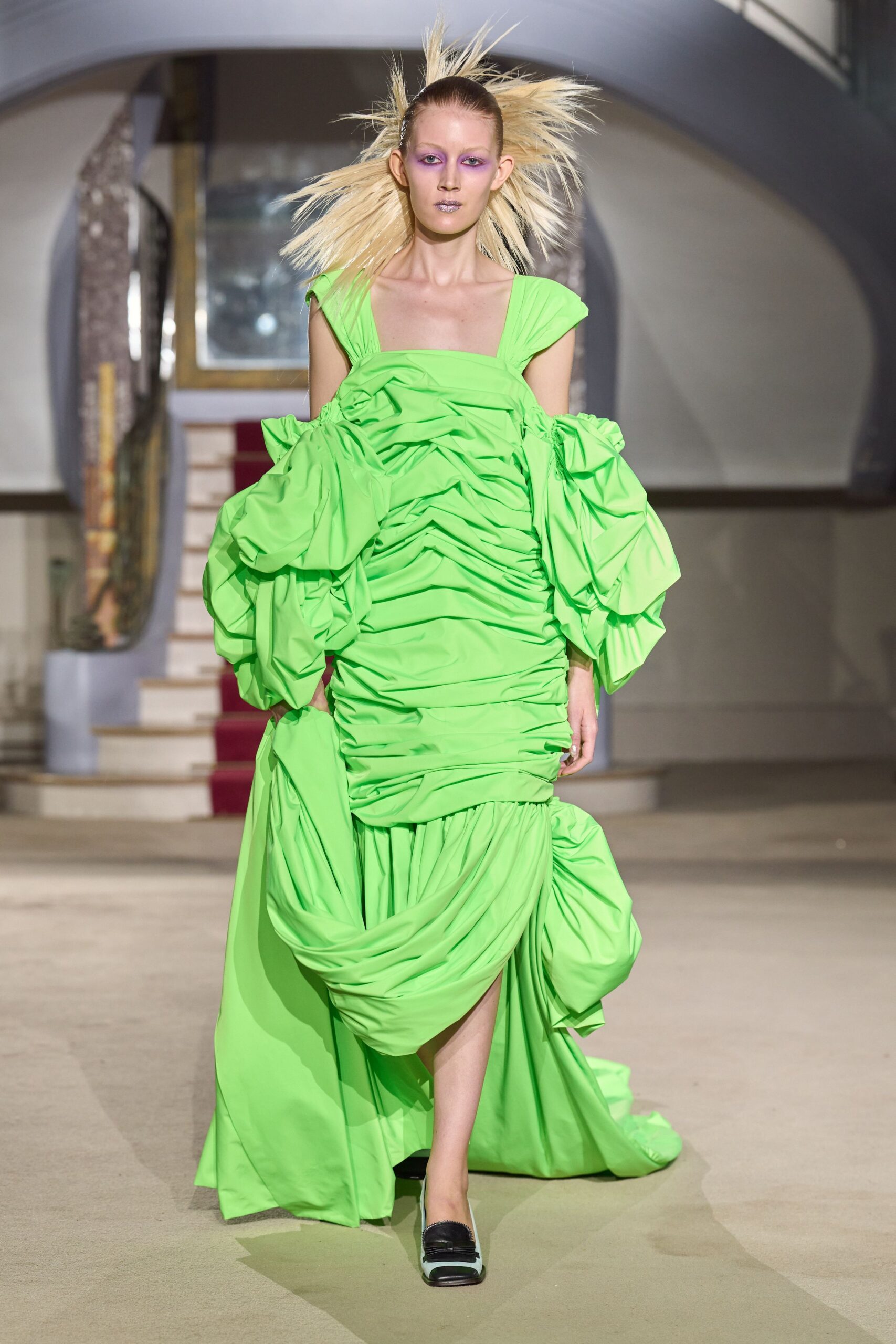 Acid-green gown in crisp taffeta, ruched tight through the torso. Off-shoulder puff sleeves ballooning at both arms. Dropped skirt swathed into heavy gathered drapes, slit opening mid-step. Black-and-white loafers with a squared vamp. Crossing a hotel atrium after midnight with flowers already crushed.