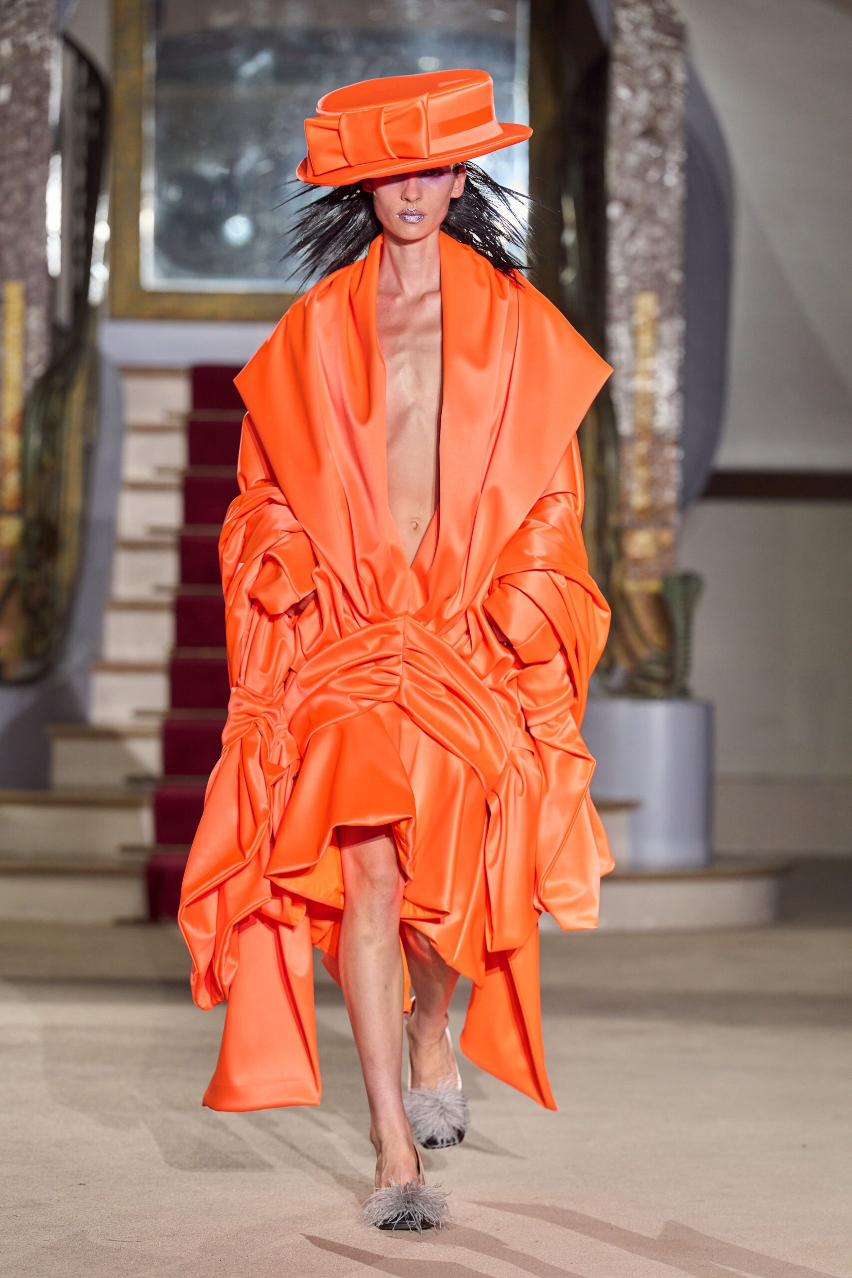 Fluorescent orange satin hat with a wide folded band. Acid-orange duchesse satin dress, ruched hard at the waist into heavy cascades. Grey feather-trimmed mules. Crossing a hotel atrium before the wedding guests have come downstairs.